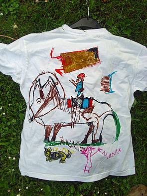 Ein von einem Kind in bunten Farben selbstbemaltes, ursprünglich weißes T-Shirt (Rückseite). Zu sehen sind ein Pferd mit Reiterin oder Reiter sowie ein Hund und eine Frau.