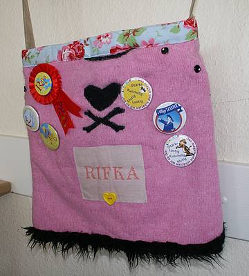Eine rosafarbene Umhängetasche, in der Mitte ist ein schwarzes Herz mit gekreuzten Knochen zu sehen. Darunter steht "Rifka". Drumherum wurden mehrere Buttons angepinnt, auf denen Rifka beschrieben wird.