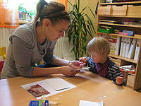 Eine Frau sitzt mit einem Jungen im Kindergartenalter am Tisch und hält ihm ein Stück Papier hin.